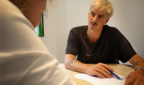 Mann mit blonden Haaren sitzt an einem Tisch und bespricht mit einem Jugendlichen einige Papiere